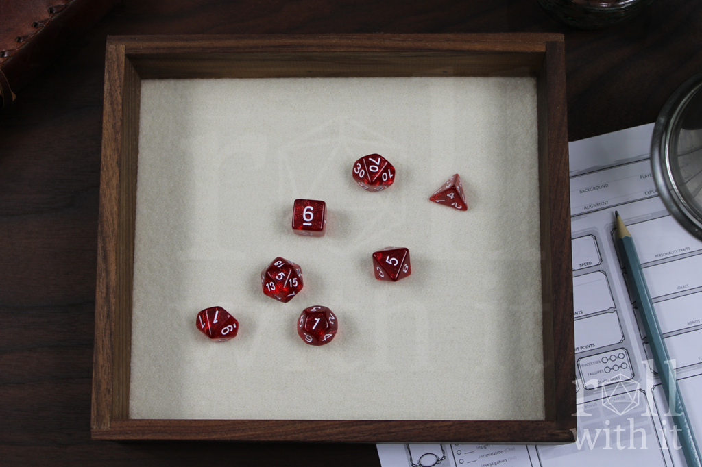 Red dice with white numbers sat on a white felt base within a solid walnut dice tray, with other tabletop accessories in the background.
