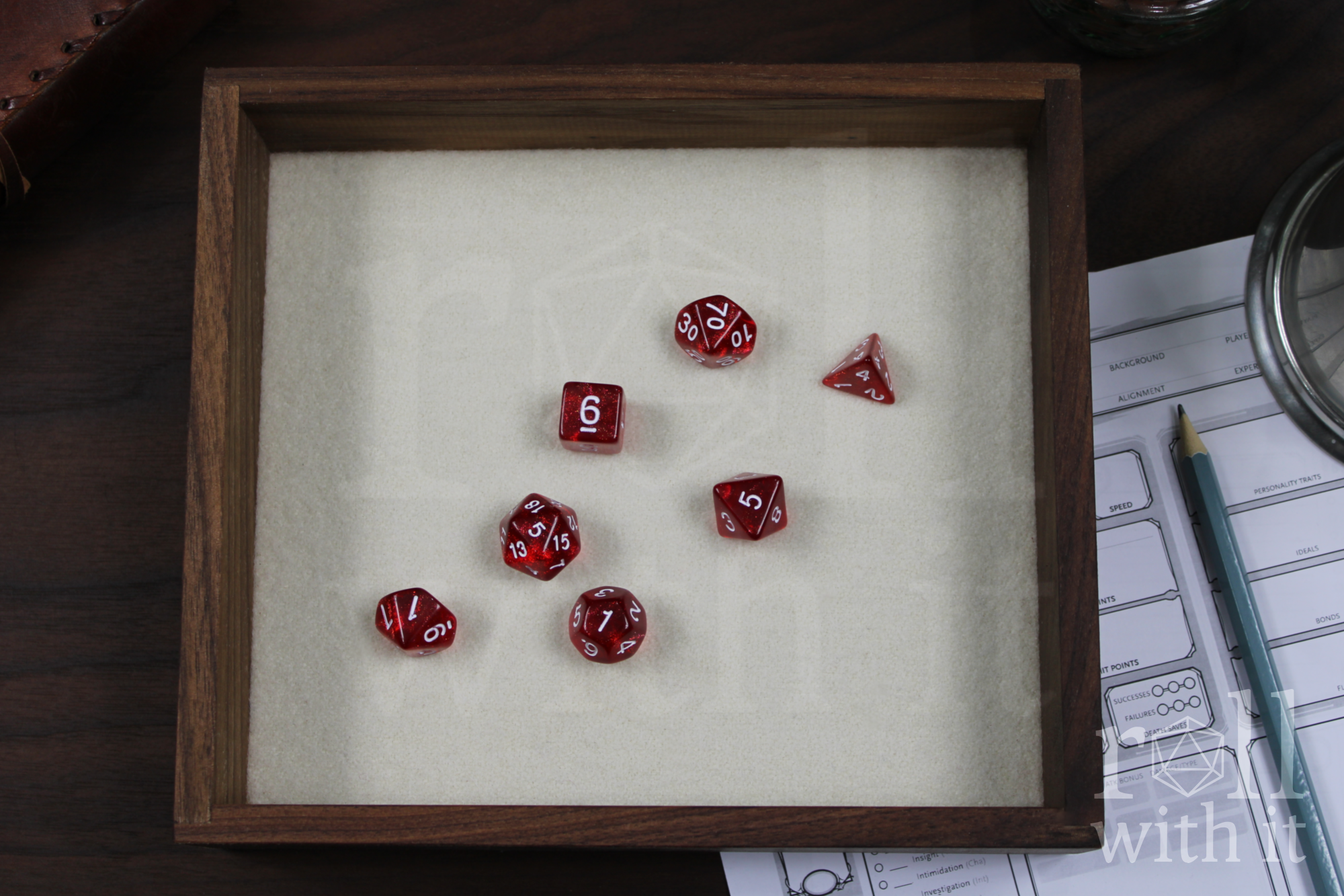 Red dice with white numbers sat on a white felt base within a solid walnut dice tray, with other tabletop accessories in the background.