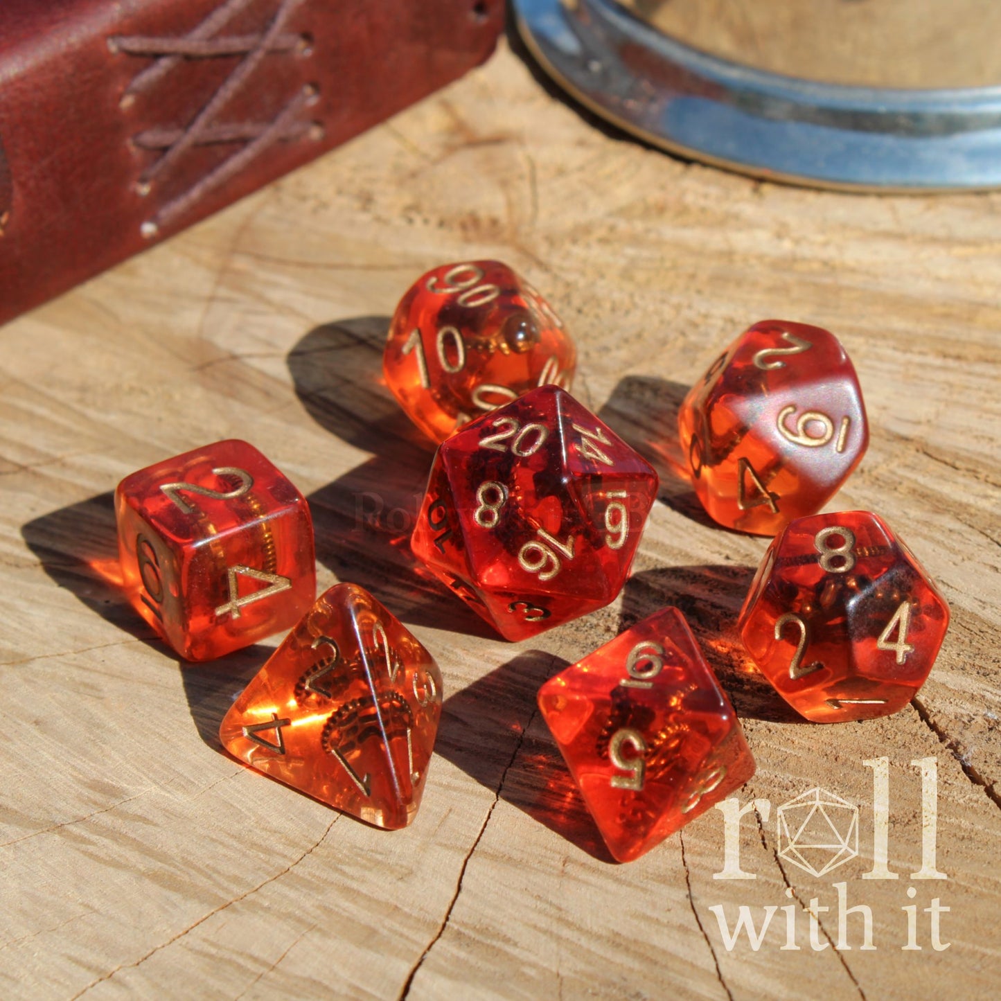 Bright shiny set of 7 red and clear polyhedral dice with golden cogs inside on a wooden surface with 'roll with it' branding.
