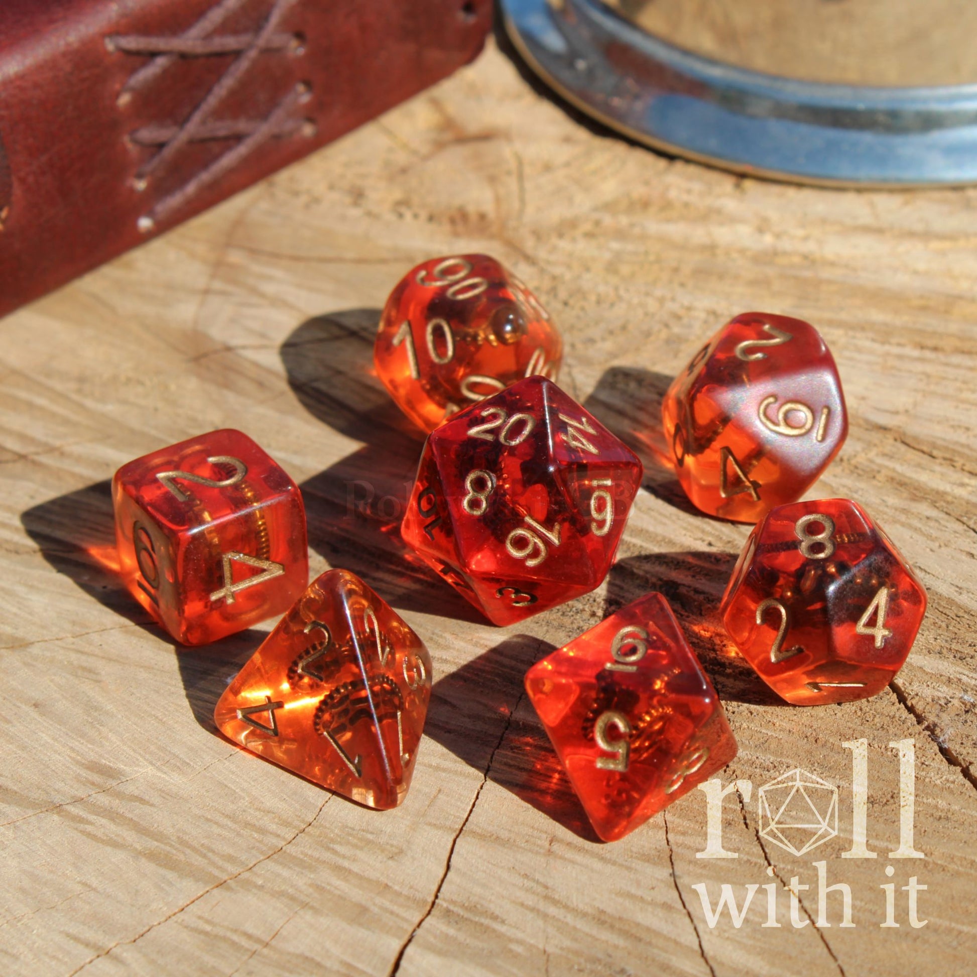 Bright shiny set of 7 red and clear polyhedral dice with golden cogs inside on a wooden surface with 'roll with it' branding.