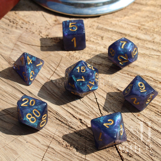 Set of 7 blue glitter filled star nebula aesthetic polyhedral dice with gold numbers on a wooden surface