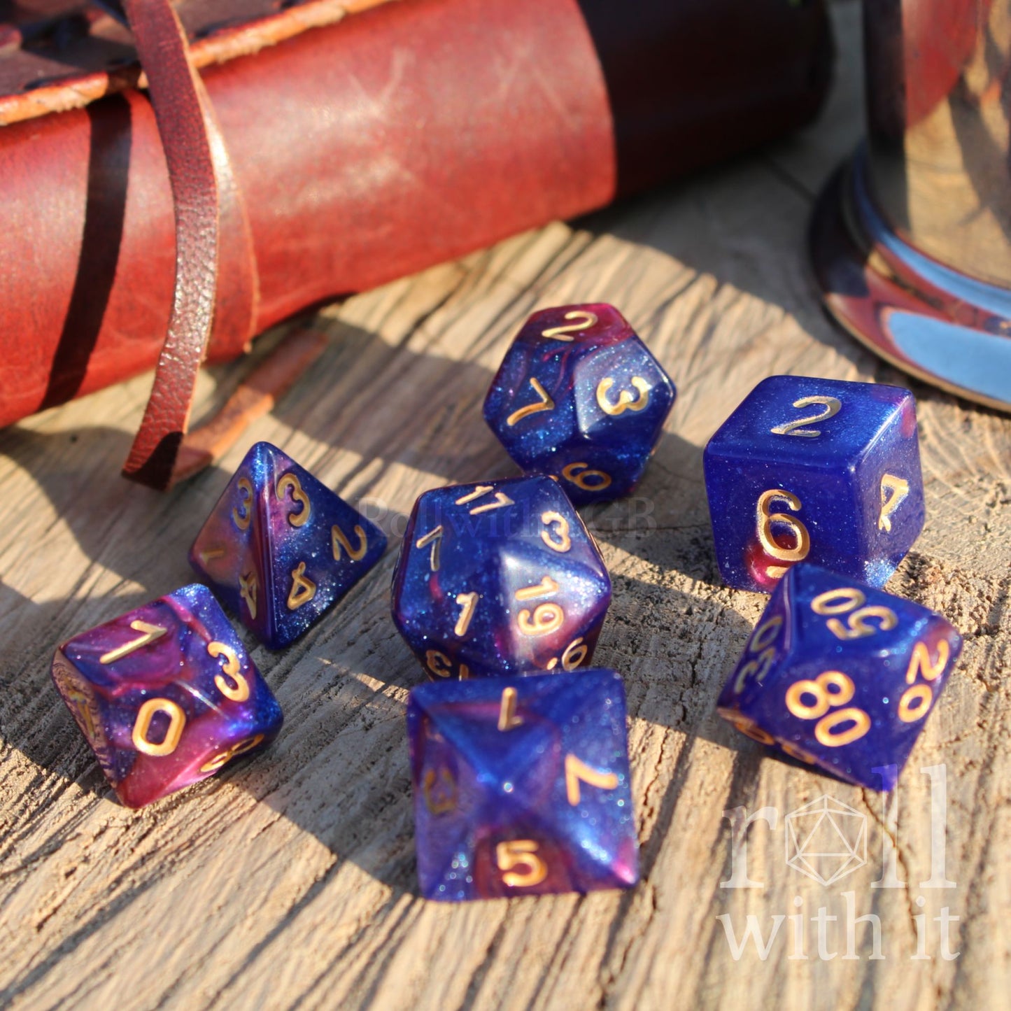 Set of blue and purple polyhedral dice with gold numbers on a wooden surface, with a leather-bound book in the background.