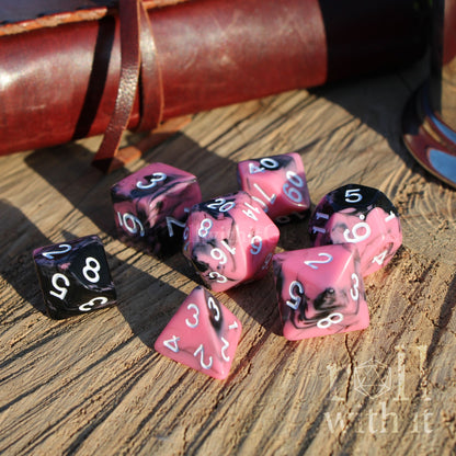 Pink and black polyhedral dice set on a wooden surface with a leather case in the background.