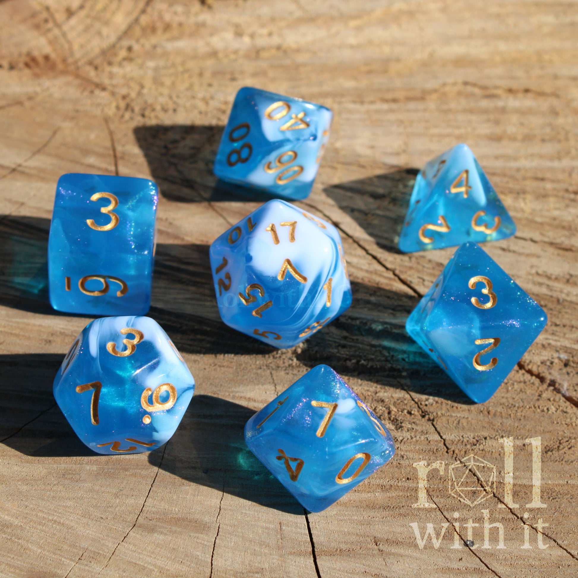 Set of bright blue and clear polyhedral dice with gold numbers on a wooden surface, featuring the text 'roll with it'.
