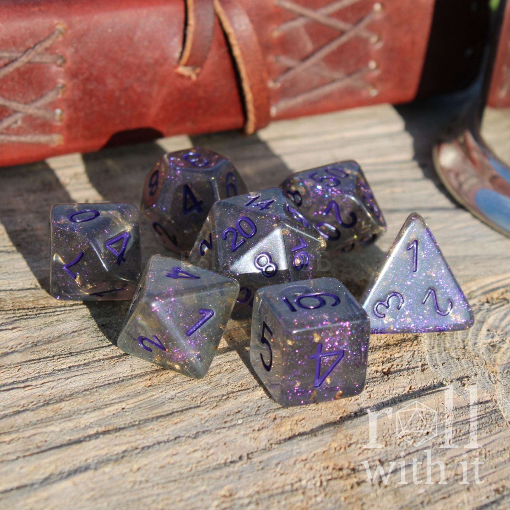 Set of purple grey polyhedral dice on a wooden surface with a leather-bound book in the background.