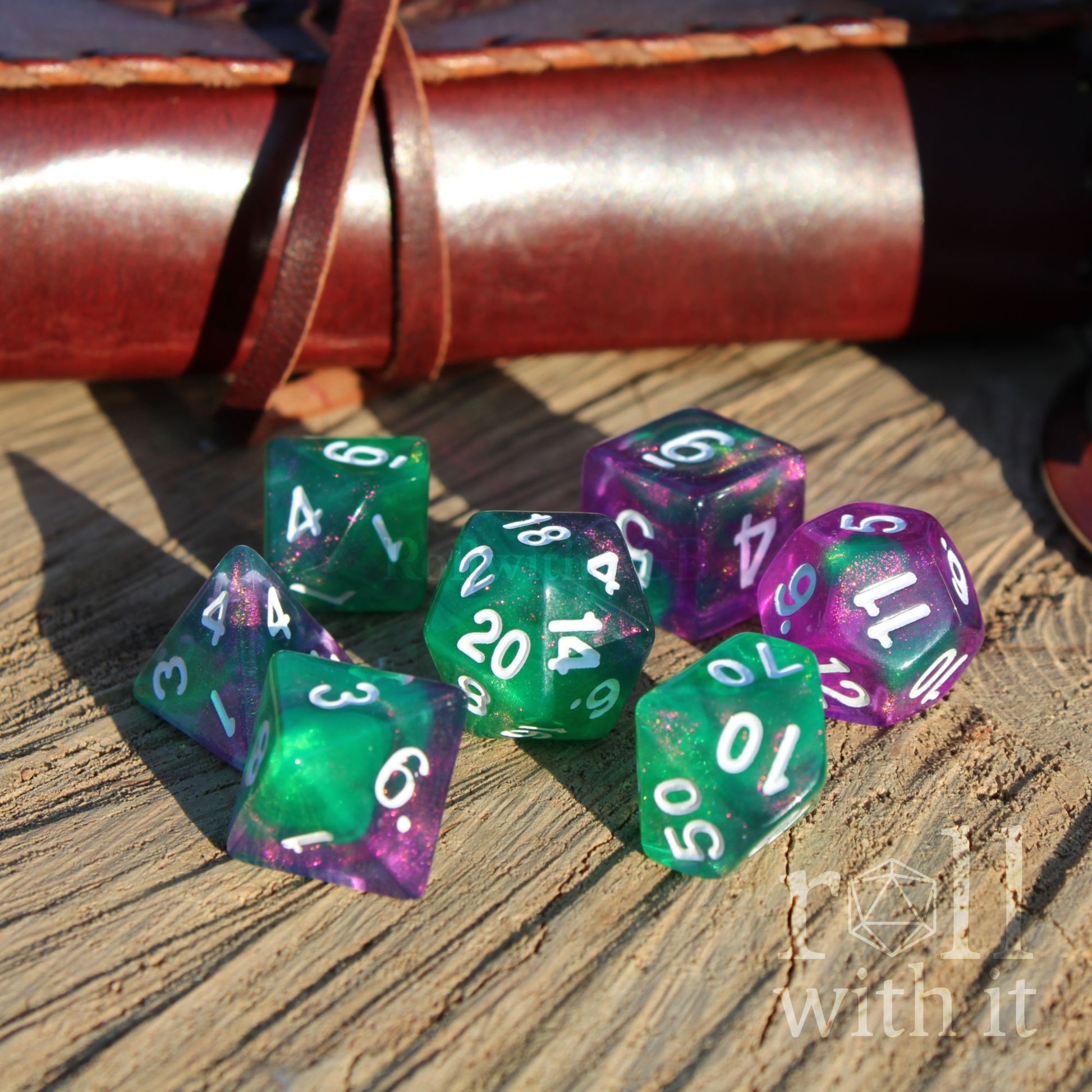 Green and purple polyhedral dice set on a wooden surface with a leather-bound book in the background.