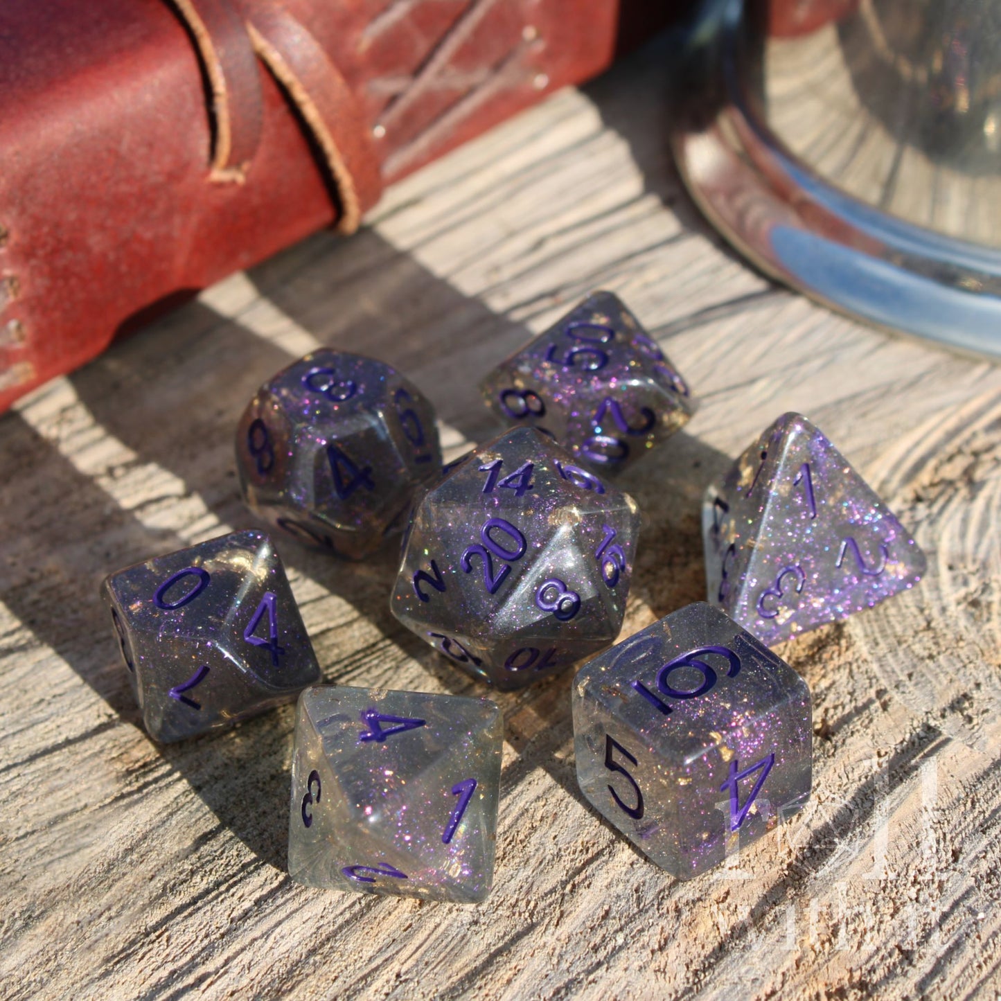 Set of purple and clear grey gold foil filled polyhedral dice on a wooden surface with a leather book in the background.