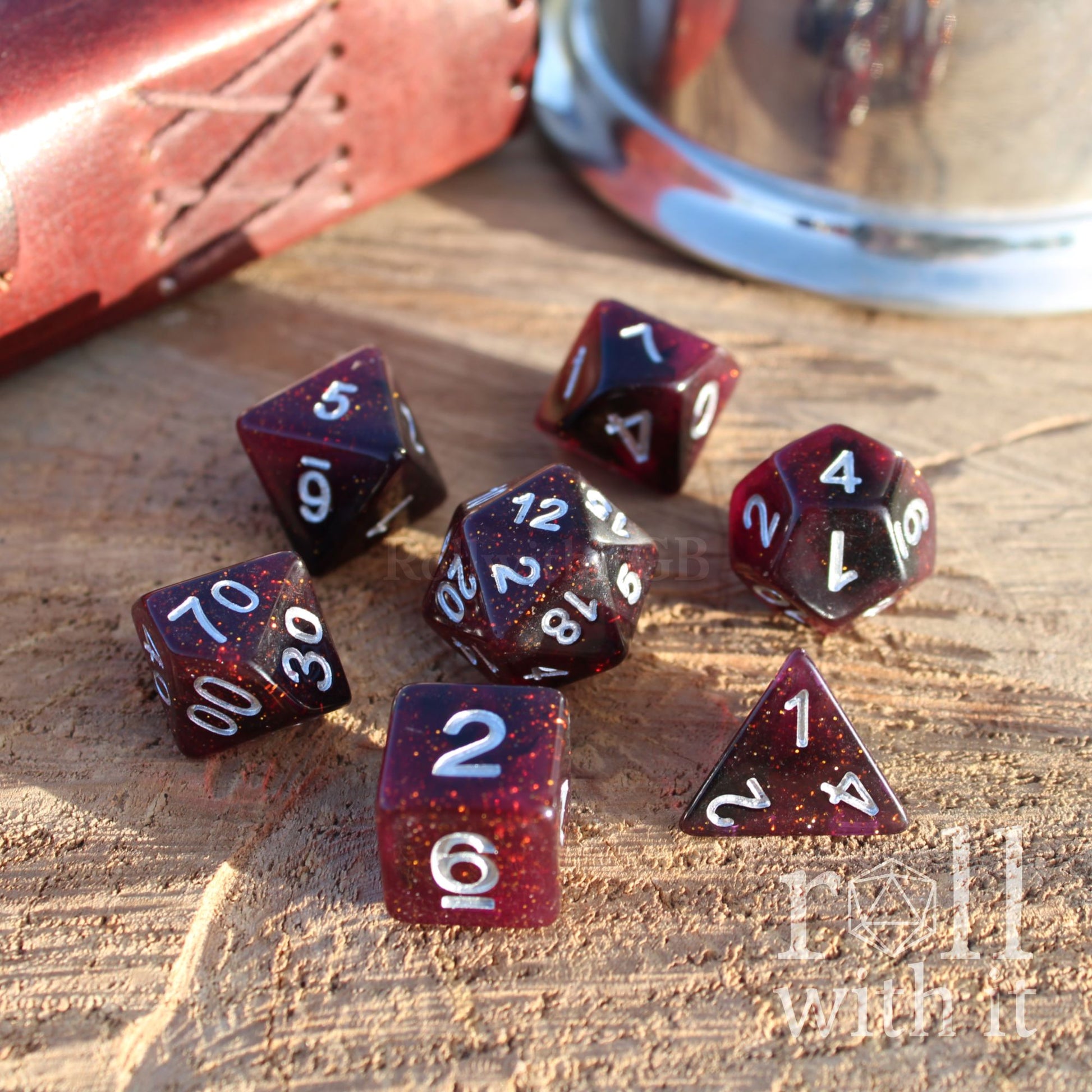 Set of maroon purple polyhedral dice with golden glitter stars on a wooden surface with a leather case and metal cup in the background.