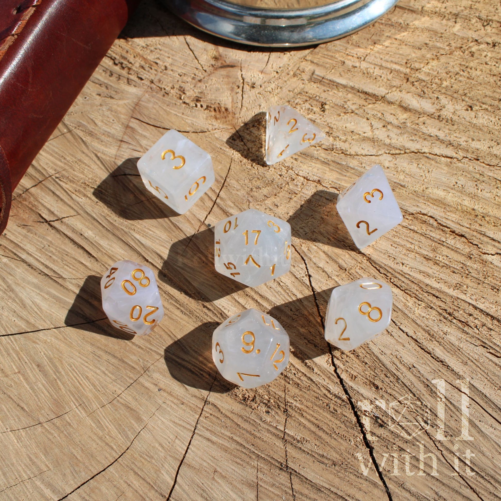 7 transparent ice white tabletop roleplaying polyhedral dice with gold numbers on a wooden surface, featuring the brand 'roll with it'.