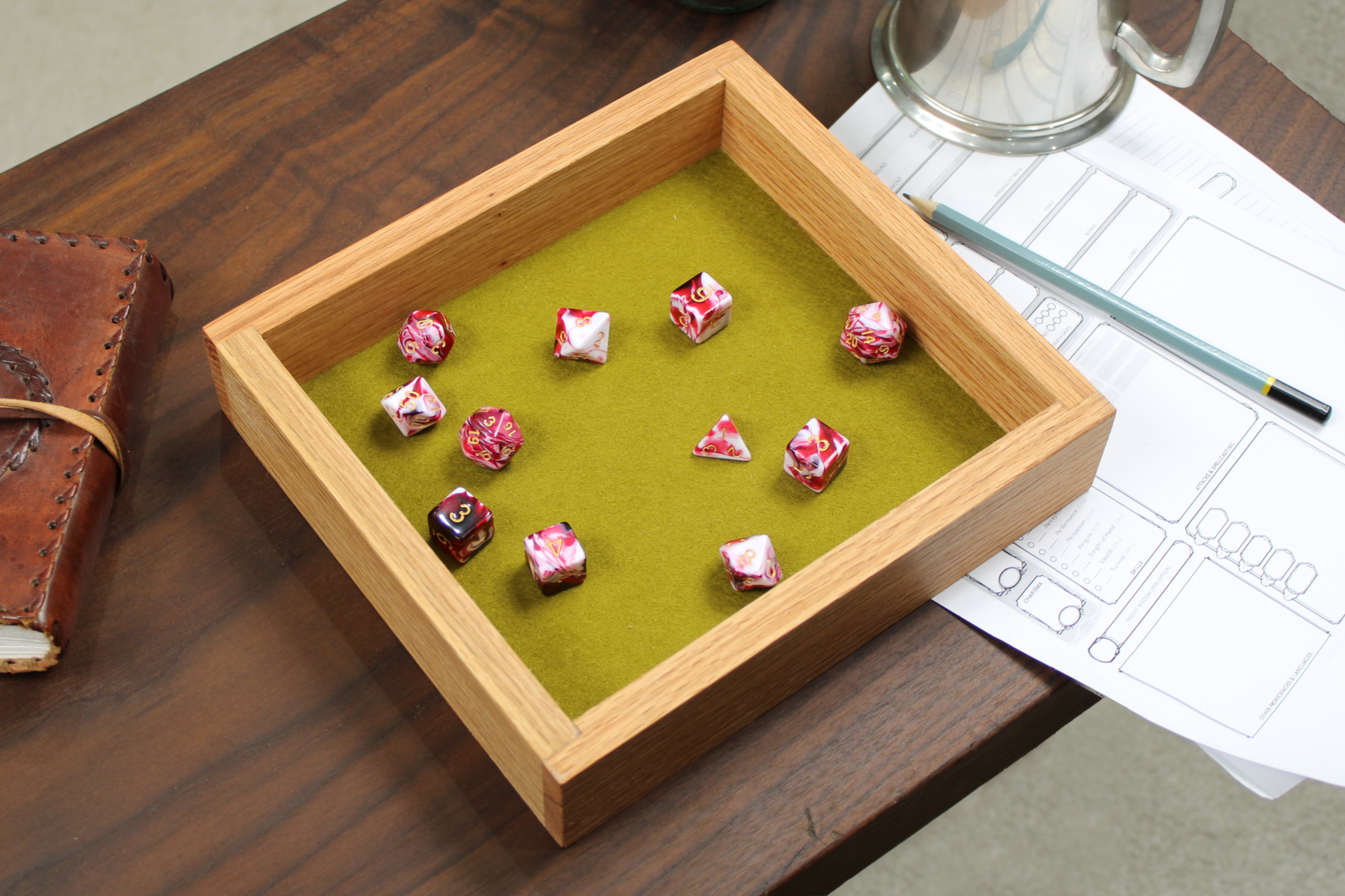 Wooden red oak role playing dice tray with green felt interior on a wooden table, featuring 'roll with it' logo. Red and cream polyhedral dice sit on the felt base.  Other tabletop accessories are in the background.