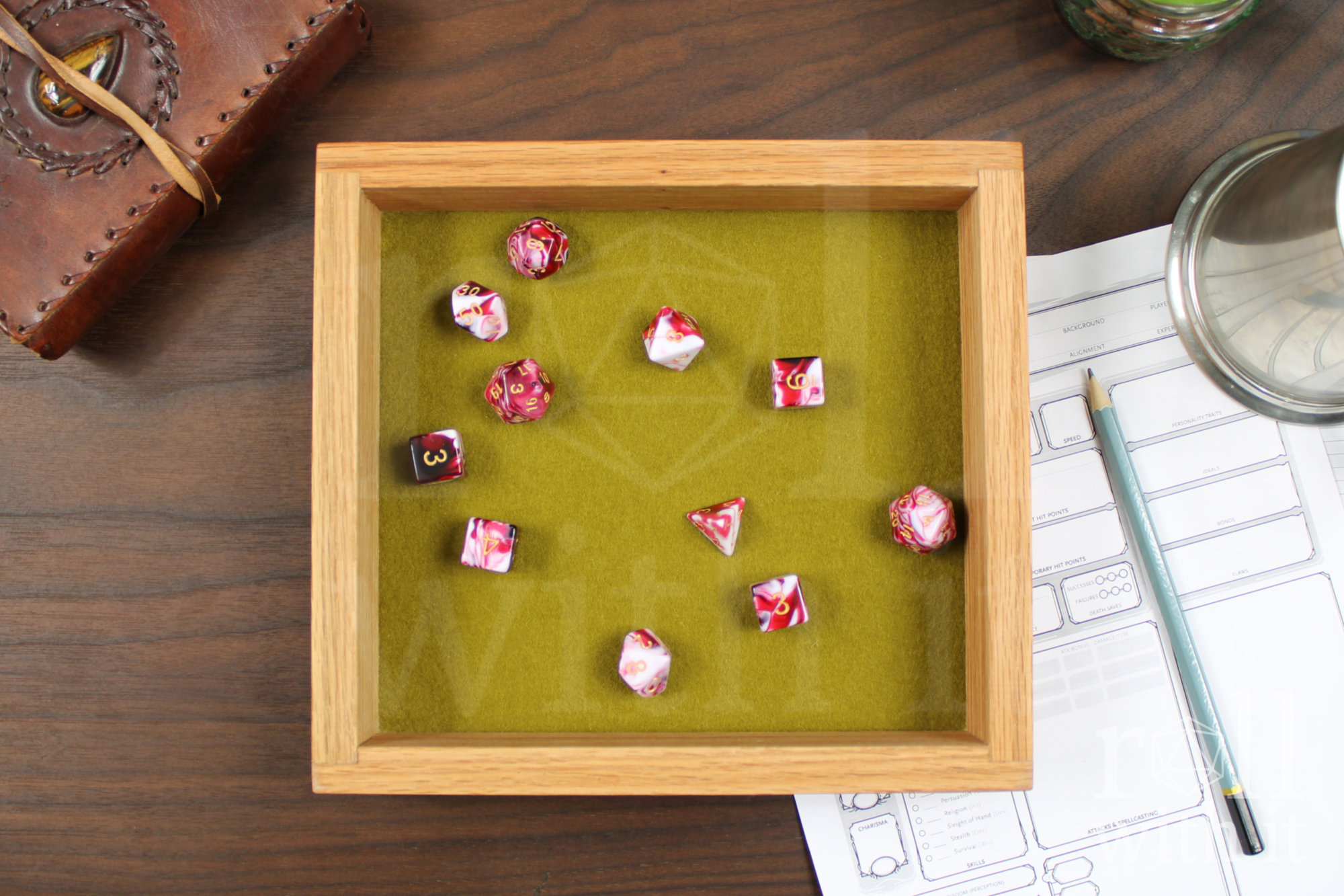 Wooden red oak dice tray with green felt interior on a wooden table, featuring 'roll with it' logo. A set of cherry cream polyhedral dice sit in the tray. A roleplaying character sheet and other tabletop accessories are in the background.