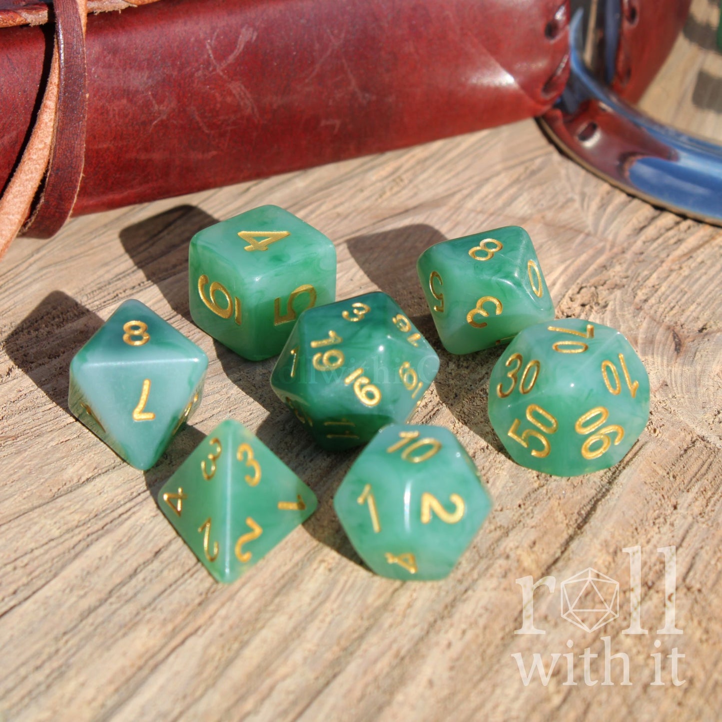 Set of green polyhedral dice with gold numbers on a wooden surface, accompanied by a leather bound book