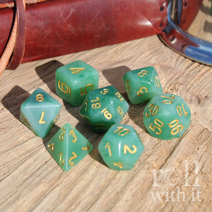 Set of green polyhedral dice with gold numbers on a wooden surface, accompanied by a leather bound book