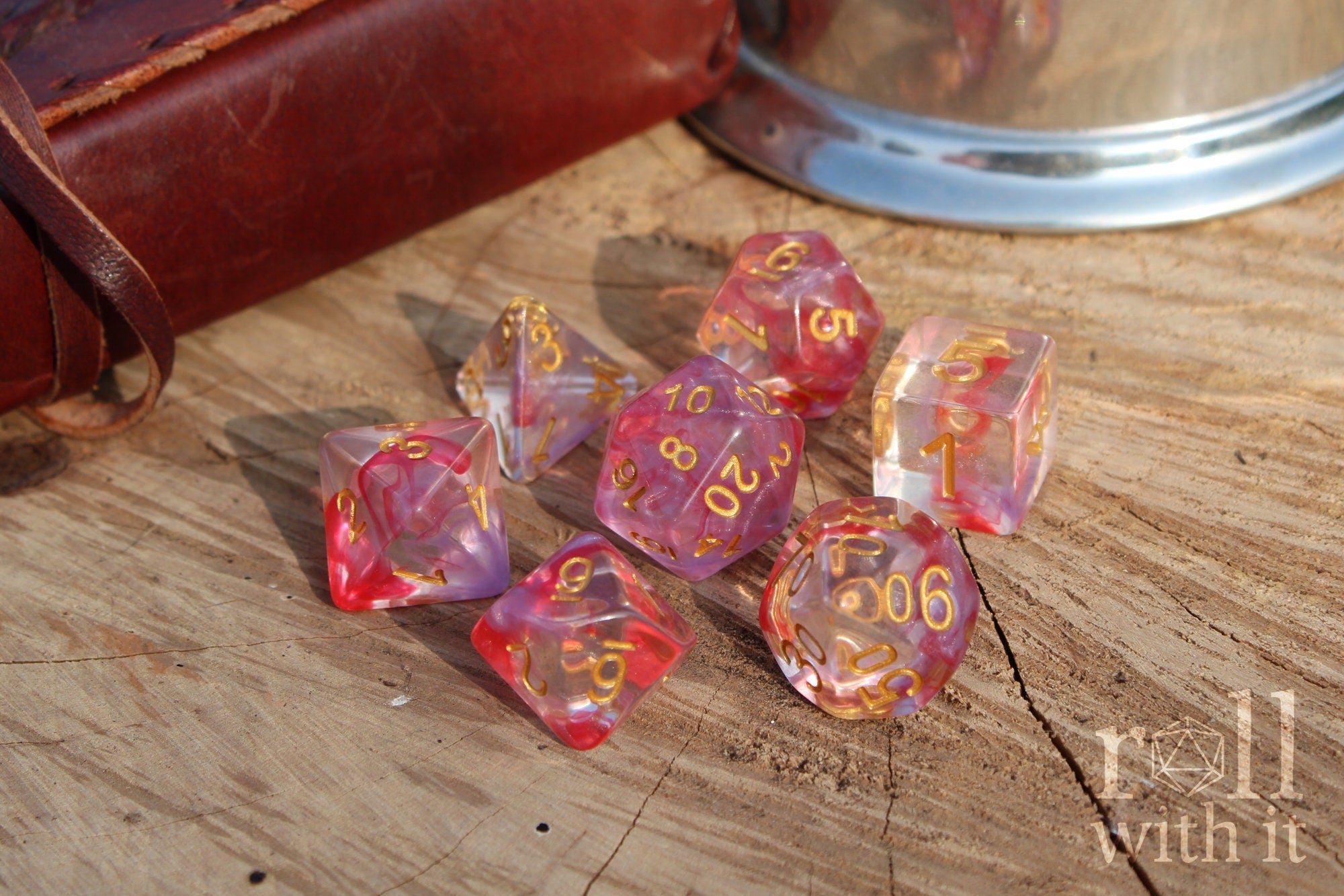 Set of translucent rose pink and purple clear roleplaying dice with gold numbers on a wooden surface with TTRPG accessories n the background