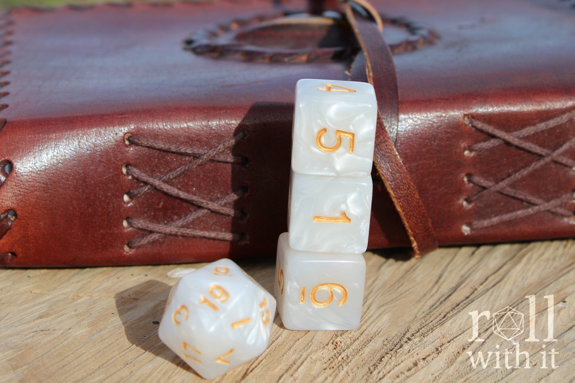 Close up of pearlescent white roleplaying DnD dice with gold numbers on a wooden surface with a brown leather journal in the background