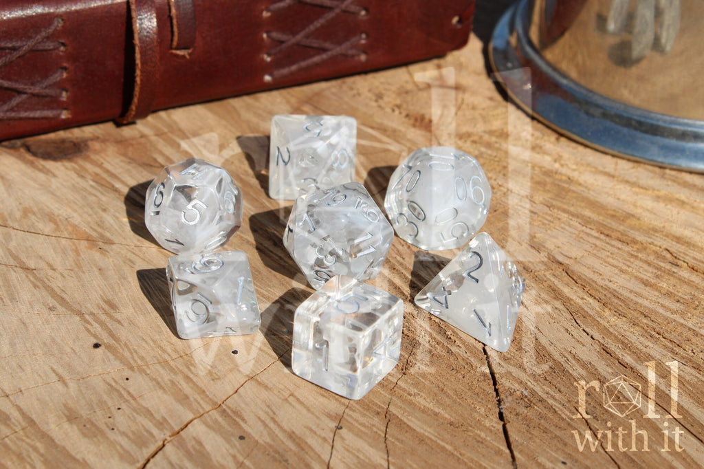 A set of 7 clear and white smoky polyhedral roleplaying dice with silver numbers on a wooden surface