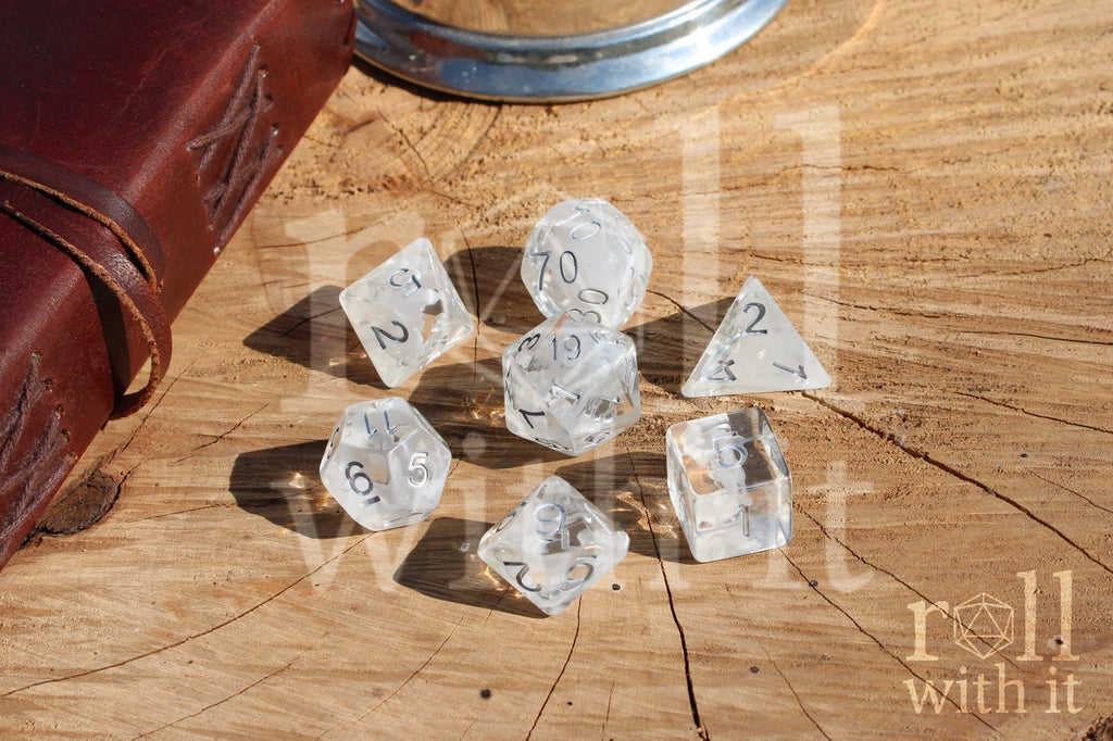 A set of 7 clear and white smoky polyhedral roleplaying dice with silver numbers on a wooden surface