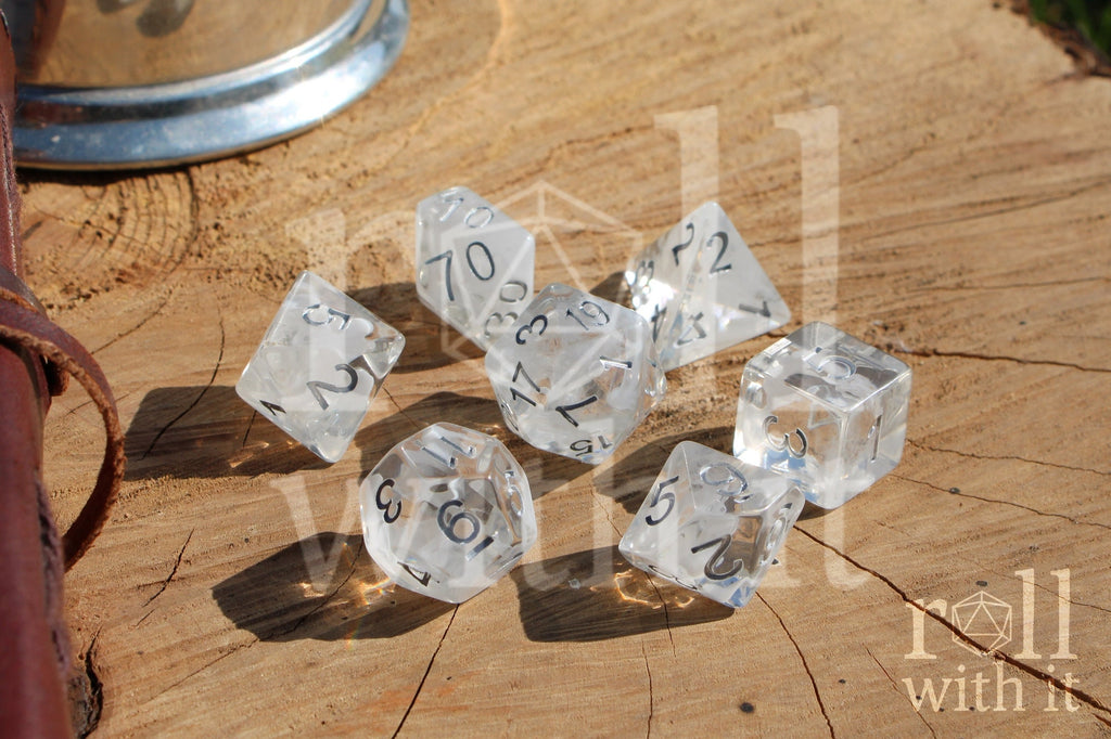 A set of 7 clear and white smoky polyhedral roleplaying dice with silver numbers on a wooden surface