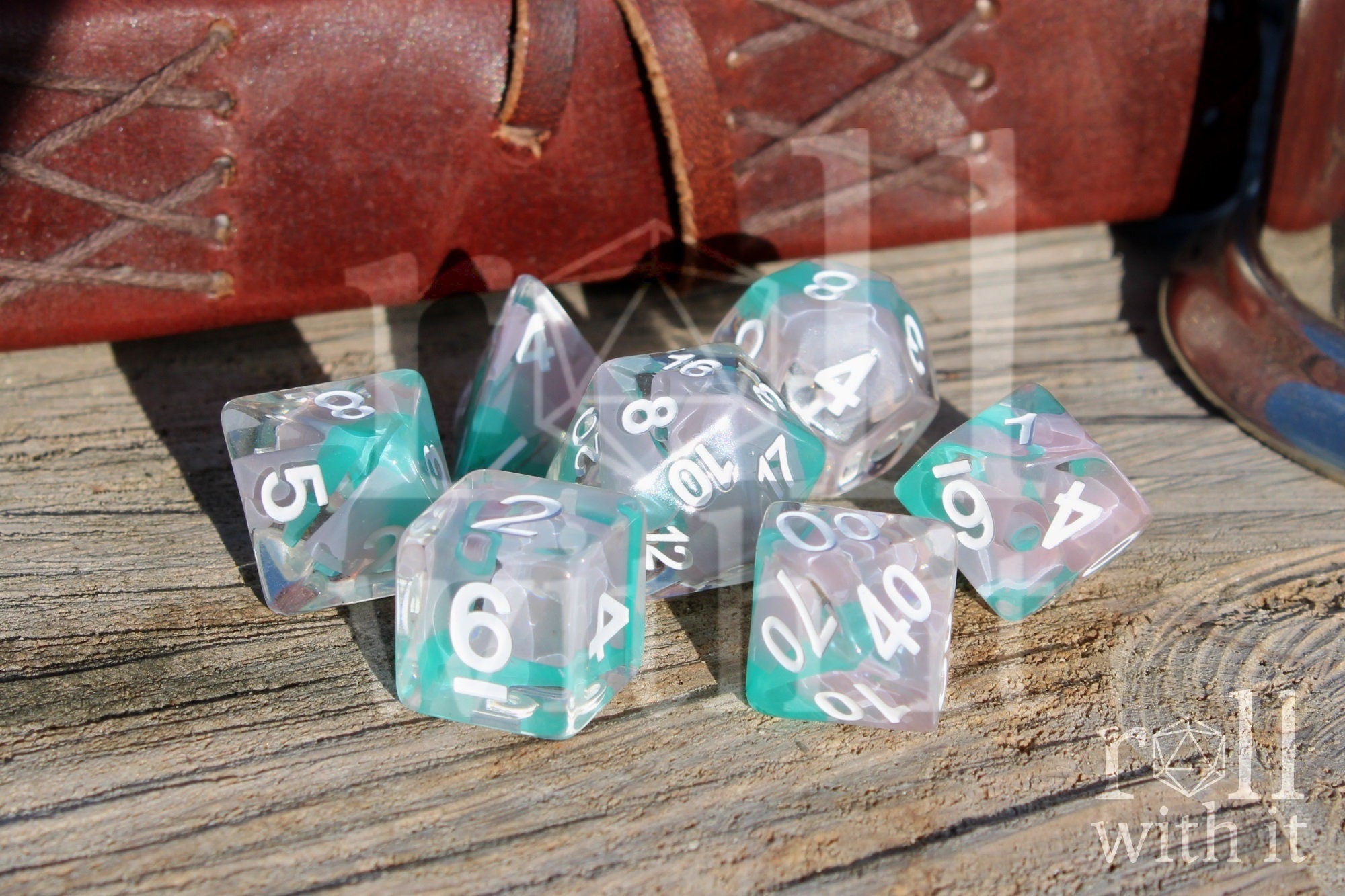 Set of light blue turquoise and white polyhedral roleplaying dice with white numbering on a wooden surface, with a brown leather bound journal in the background