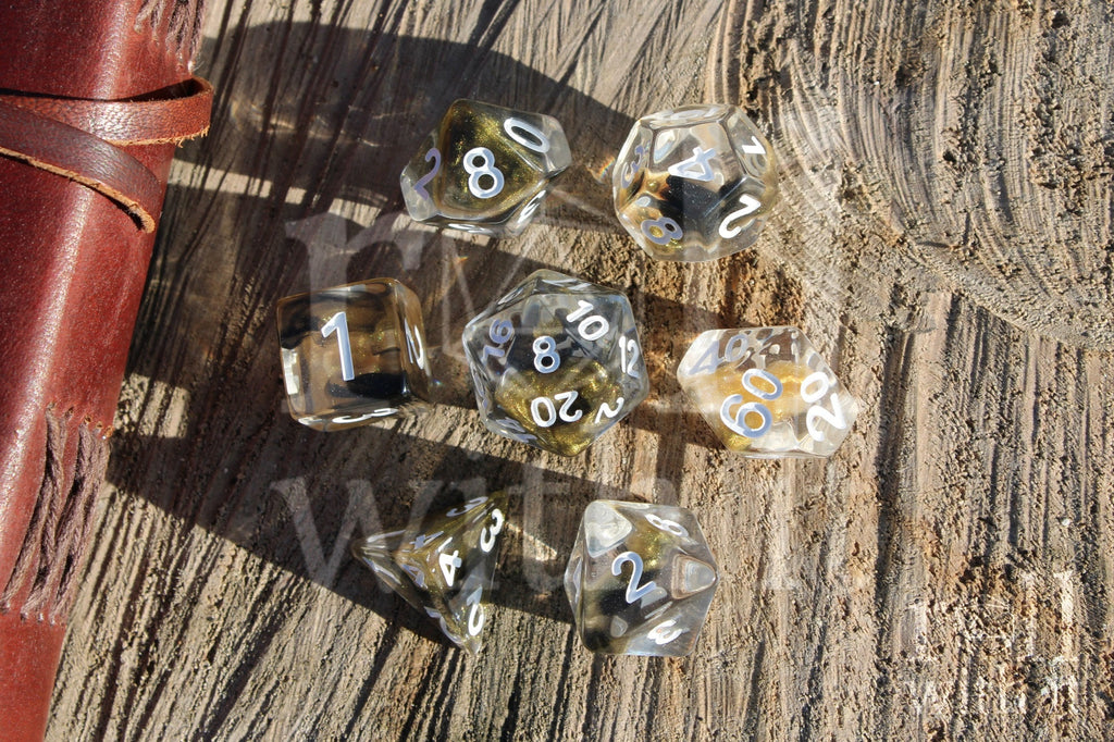 Set of 7 clear polyhedral DND dice with gold and black glittering centres, with white numbers on a wooden surface
