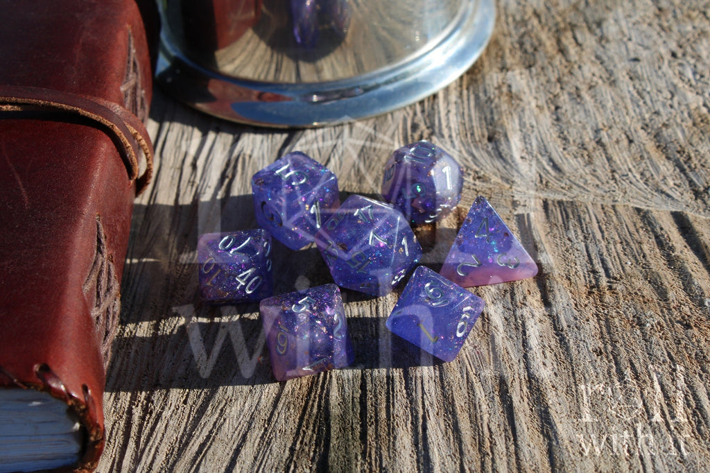 Set of purple and pink glittery and gold leaf polyhedral dice with silver lettering on a wooden surface