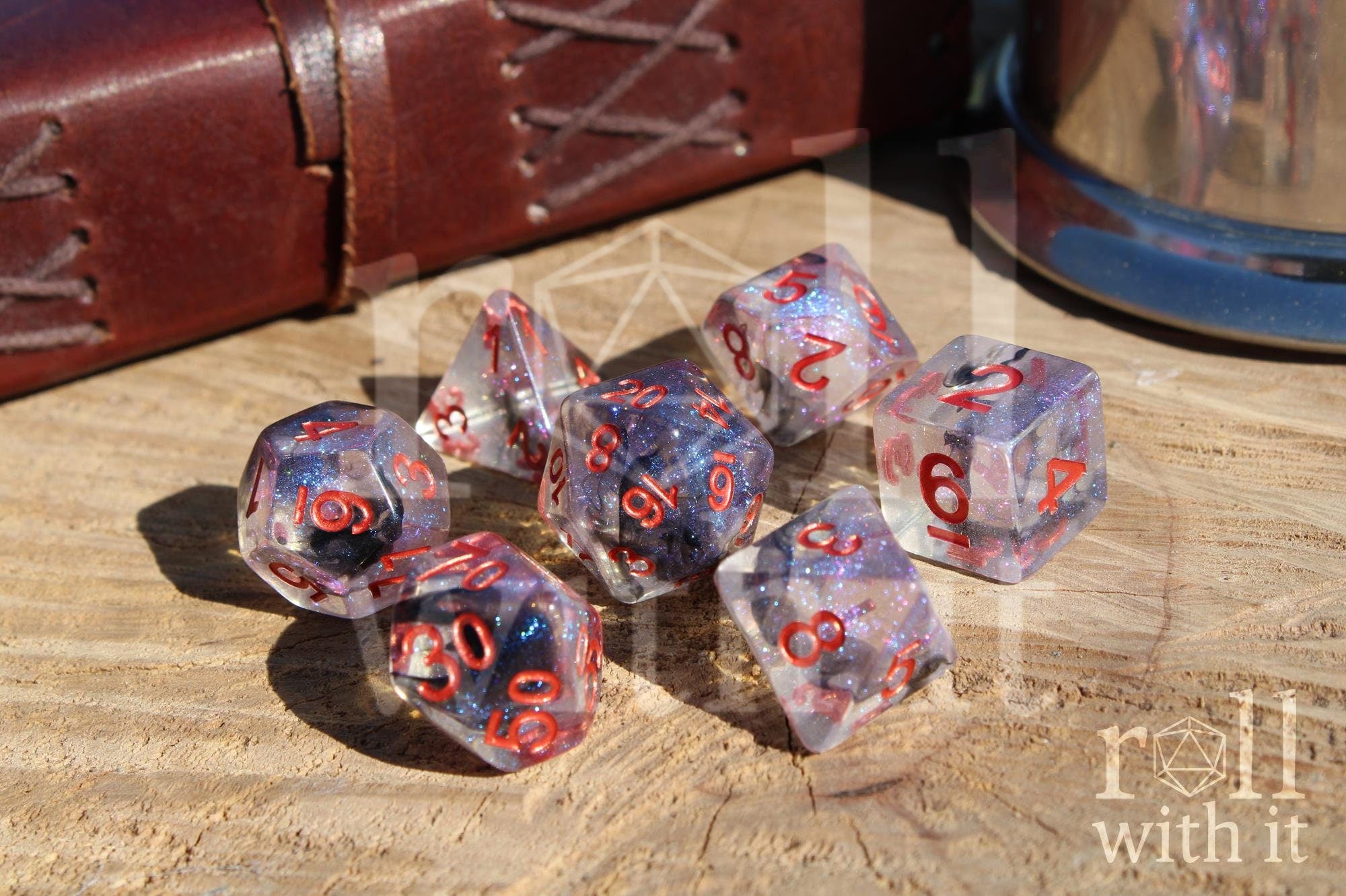 Transparent starry glittery purple and black polyhedral roleplaying dice with red numbers on a wooden surface and TTRPG accessories in the background