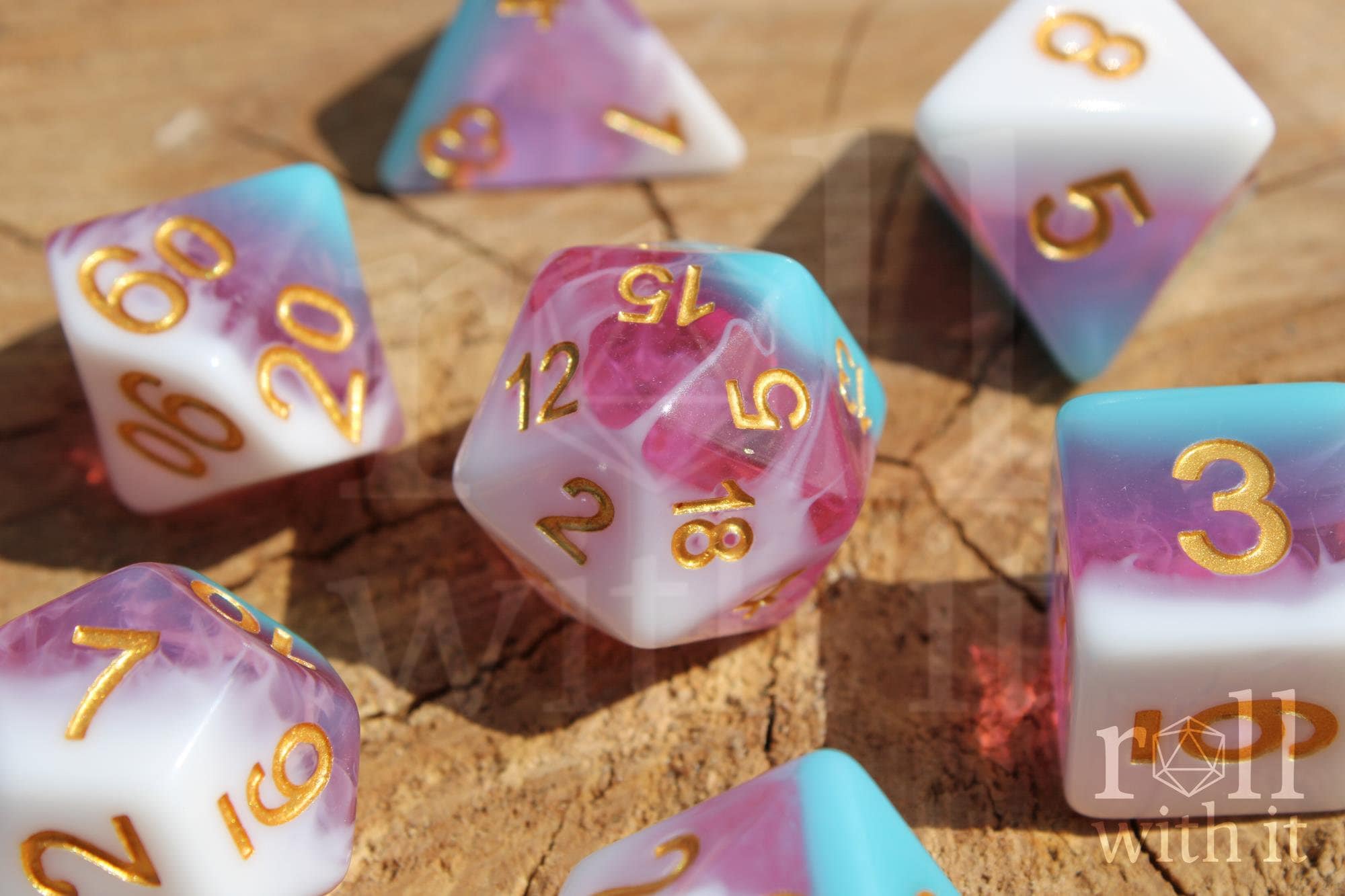 Set of colourful layered pink, light blue, and creamy white polyhedral roleplaying dice with gold numbers on a wooden surface
