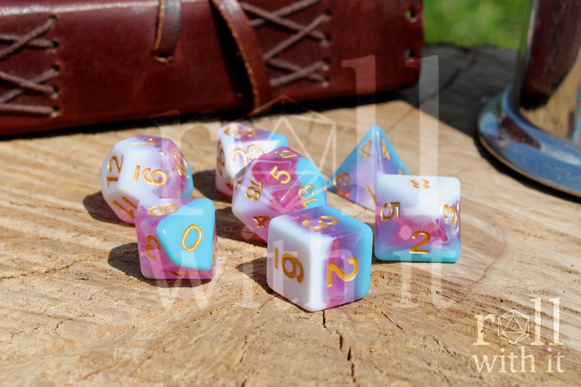 Set of colourful layered pink, light blue, and creamy white polyhedral roleplaying dice with gold numbers on a wooden surface