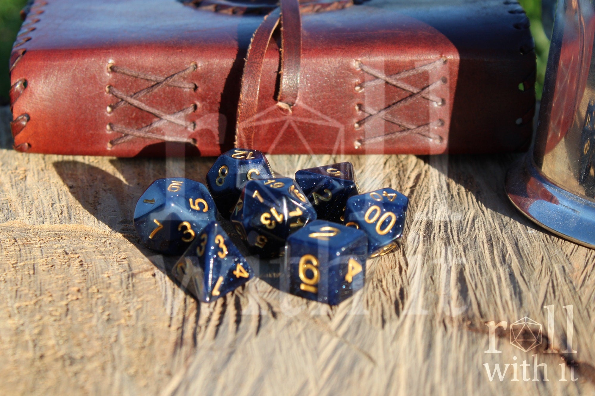 Deep midnight blue polyhedral starry glittery dice with gold numbers on a wooden surface with a leather bound journal in the background 