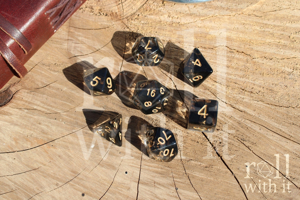 Set of clear and smoky black polyhedral roleplaying dice on a wooden surface with tabletop RPG accessories in the background