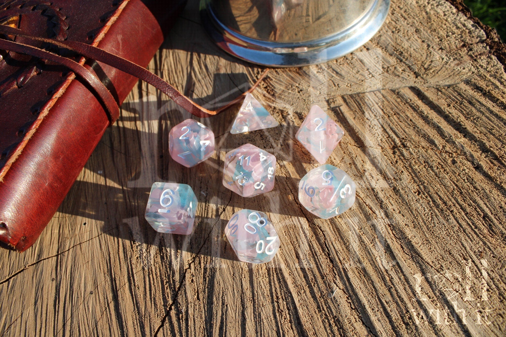 Set of pink and pale blue translucent polyhedral DND roleplaying dice on a wooden surface with a leather journal and silver tankard in the background