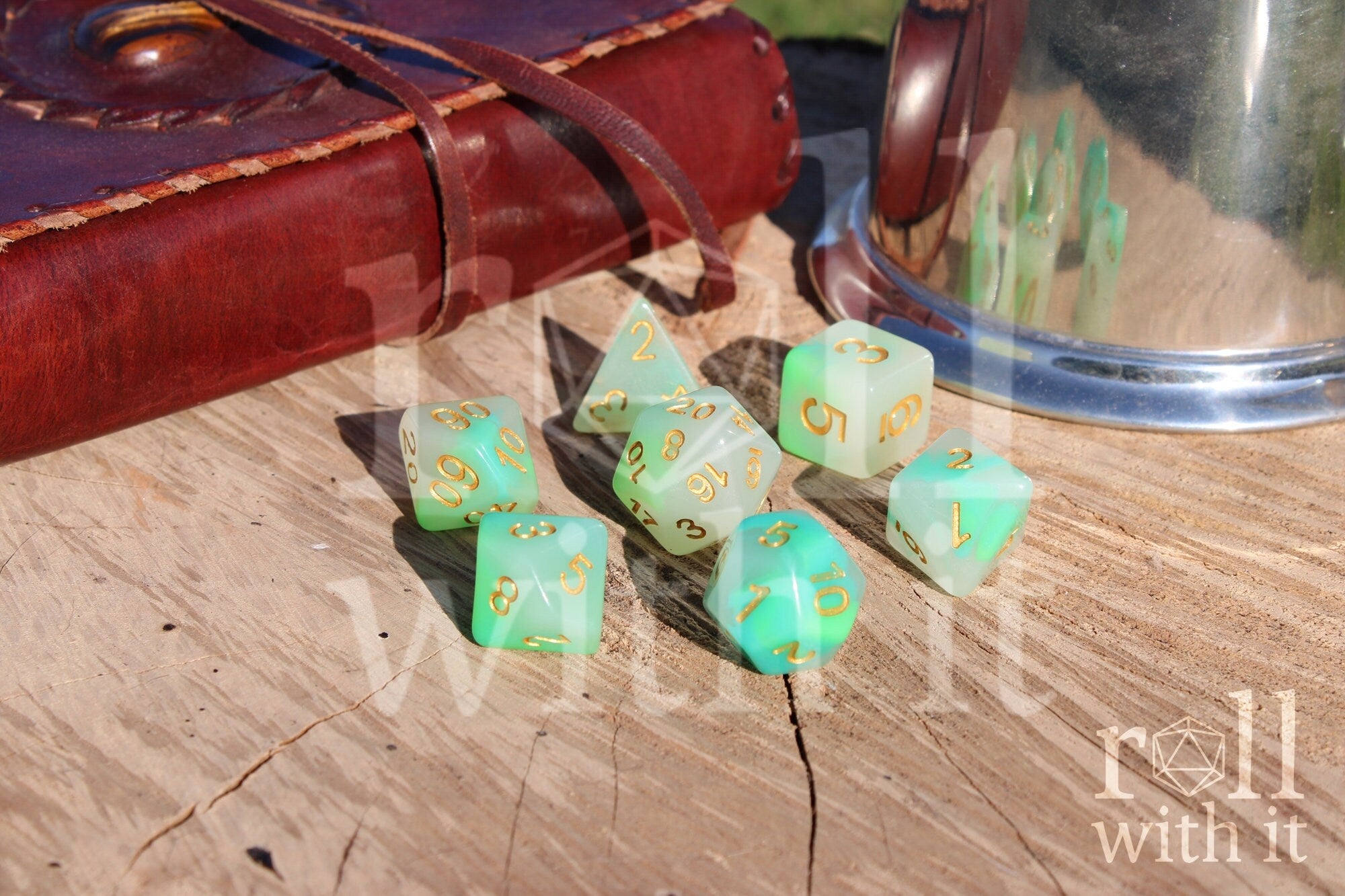 Set of turquoise and green polyhedral DND dice with gold numbers on a wooden surface with a leather journal and pewter tankard in the background.