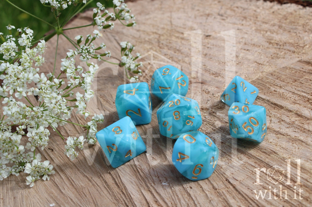 Aqua polyhedral roleplaying dice set with gold numbers on a wooden surface with white flowers.