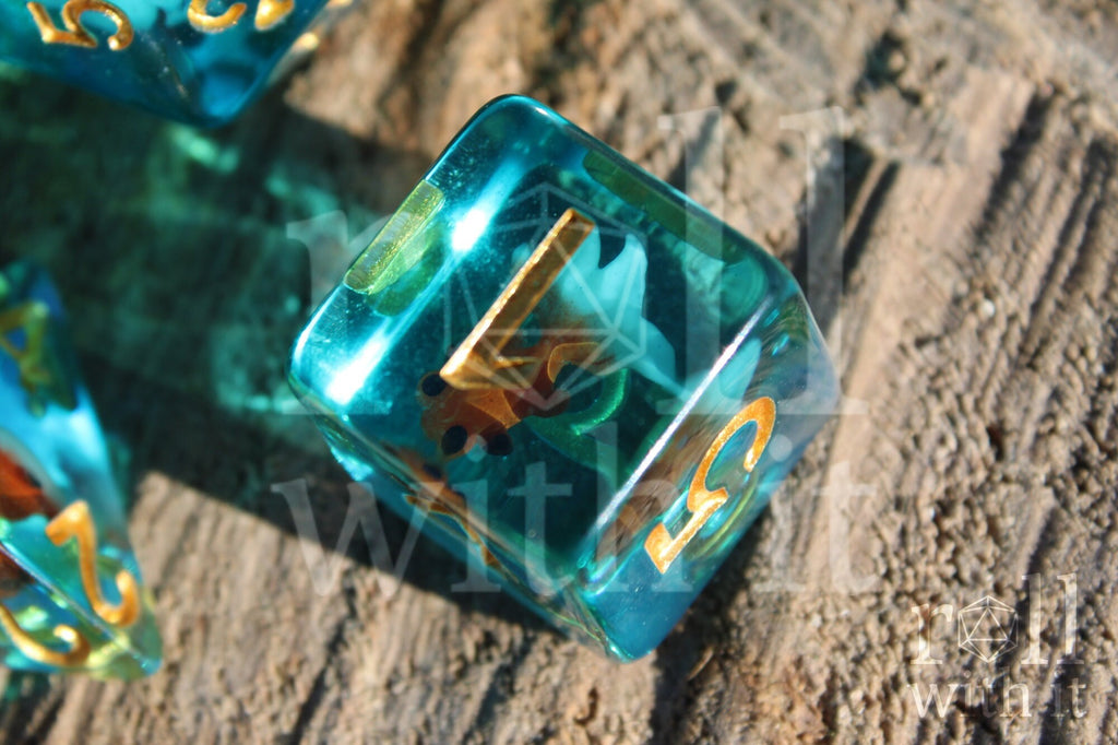 Set of clear blue turquoise polyhedral dice with gold numbers and a mini goldfish inside on a wooden surface