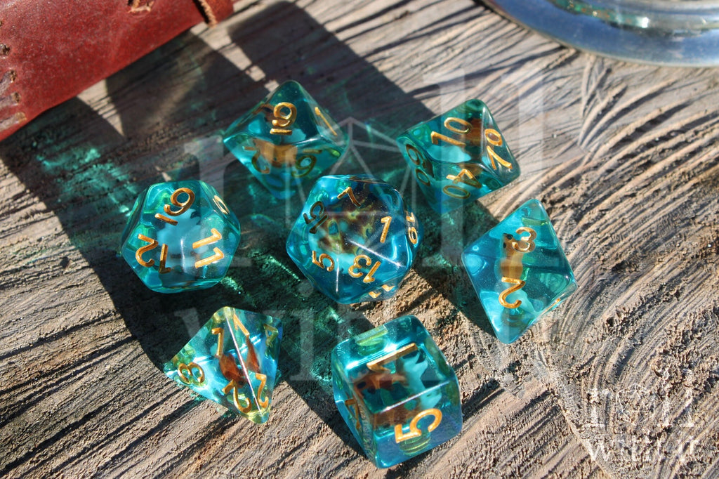 Set of clear blue turquoise polyhedral dice with gold numbers and a mini goldfish inside on a wooden surface