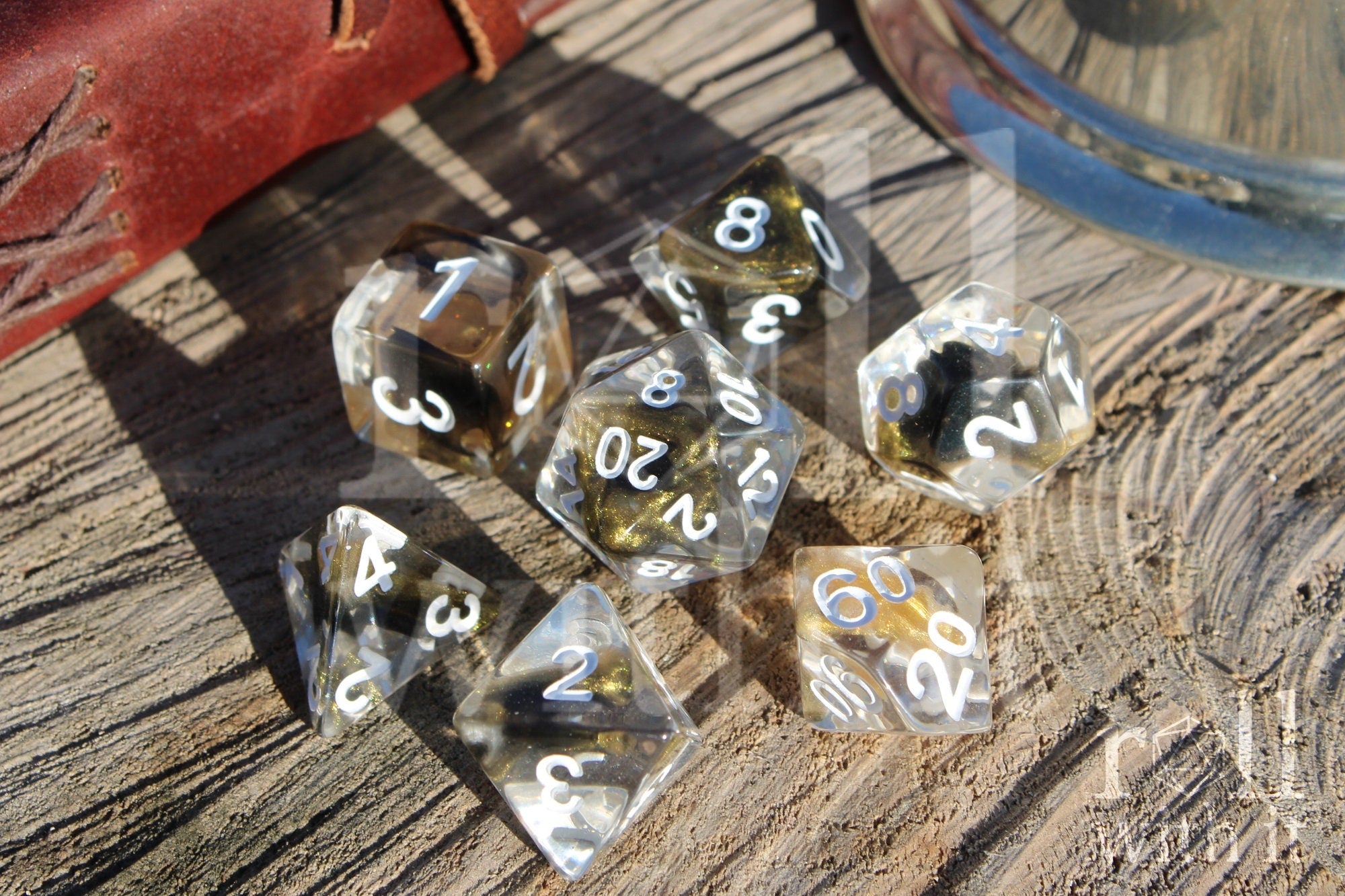 Set of 7 clear polyhedral DND dice with gold and black glittering centres, with white numbers on a wooden surface