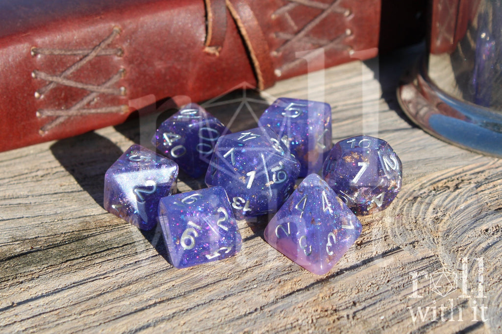 Set of purple and pink glittery and gold leaf polyhedral DND dice with silver lettering on a wooden surface
