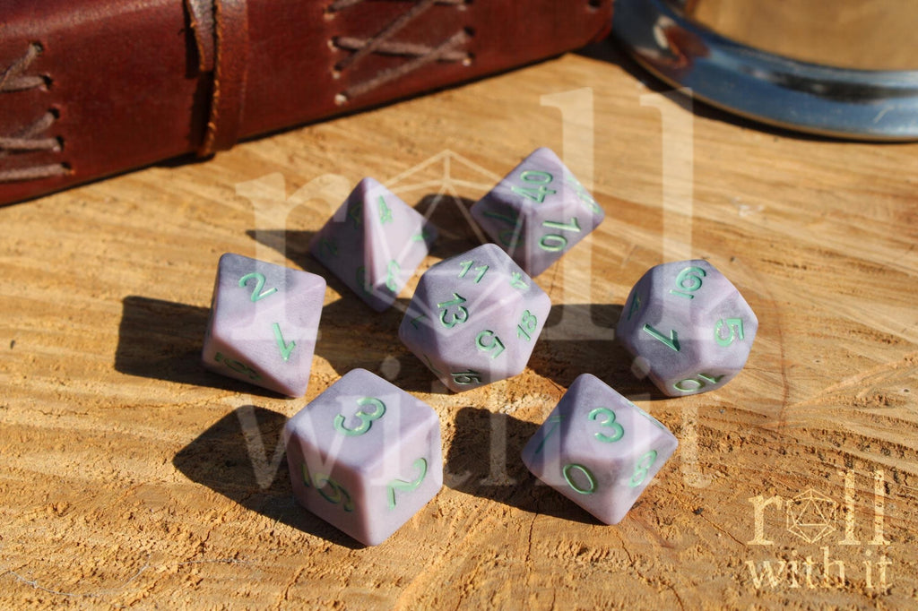 Lilac purple matte polyhedral DnD roleplaying dice with mint green numbers on a wooden surface with a brown leather journal in the background