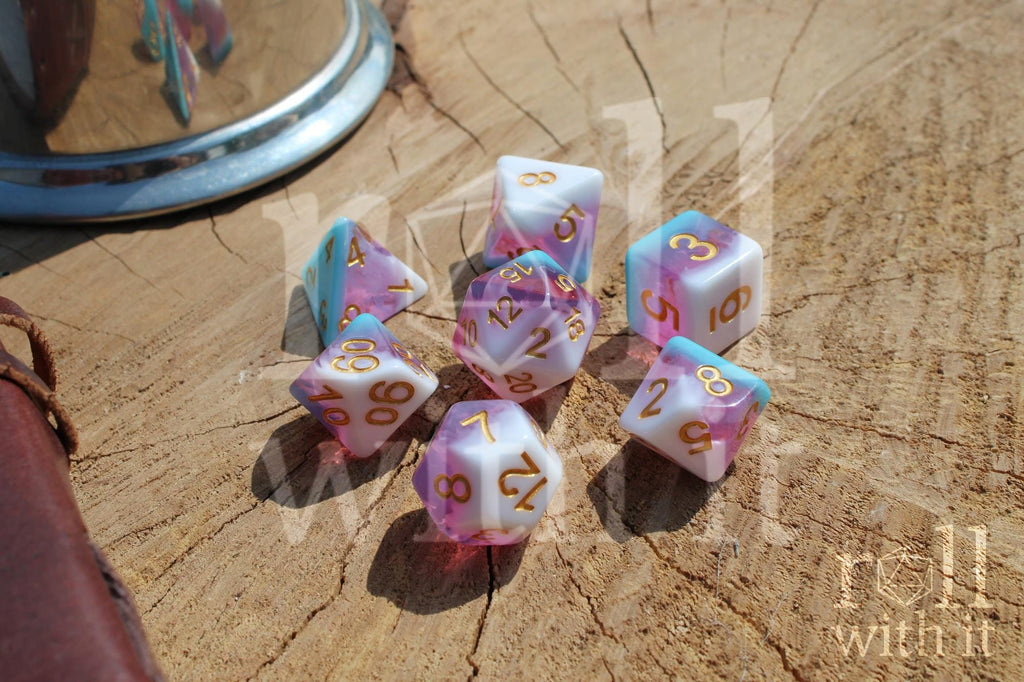 Set of colourful layered pink, light blue, and creamy white polyhedral TTRPG dice with gold numbers on a wooden surface