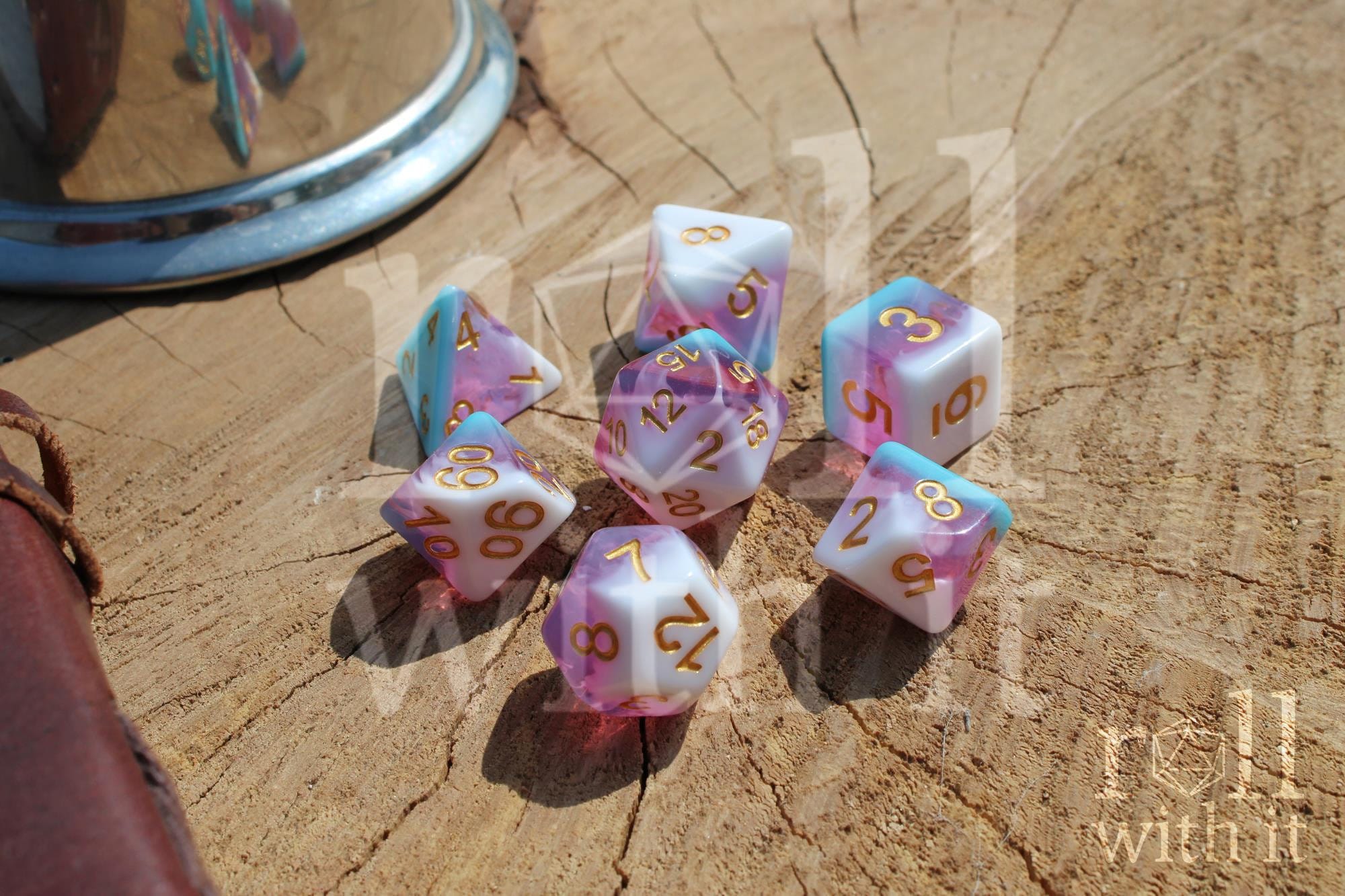 Set of colourful layered pink, light blue, and creamy white polyhedral TTRPG dice with gold numbers on a wooden surface