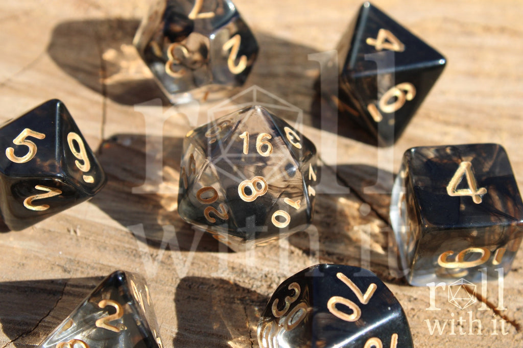 Close up of a set of clear and smoky black polyhedral roleplaying dice on a wooden surface with tabletop RPG accessories in the background