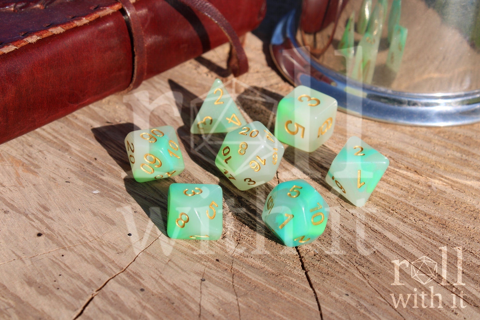 Set of turquoise and green polyhedral DND dice with gold numbers on a wooden surface with a leather bound journal and shiny tankard in the background 