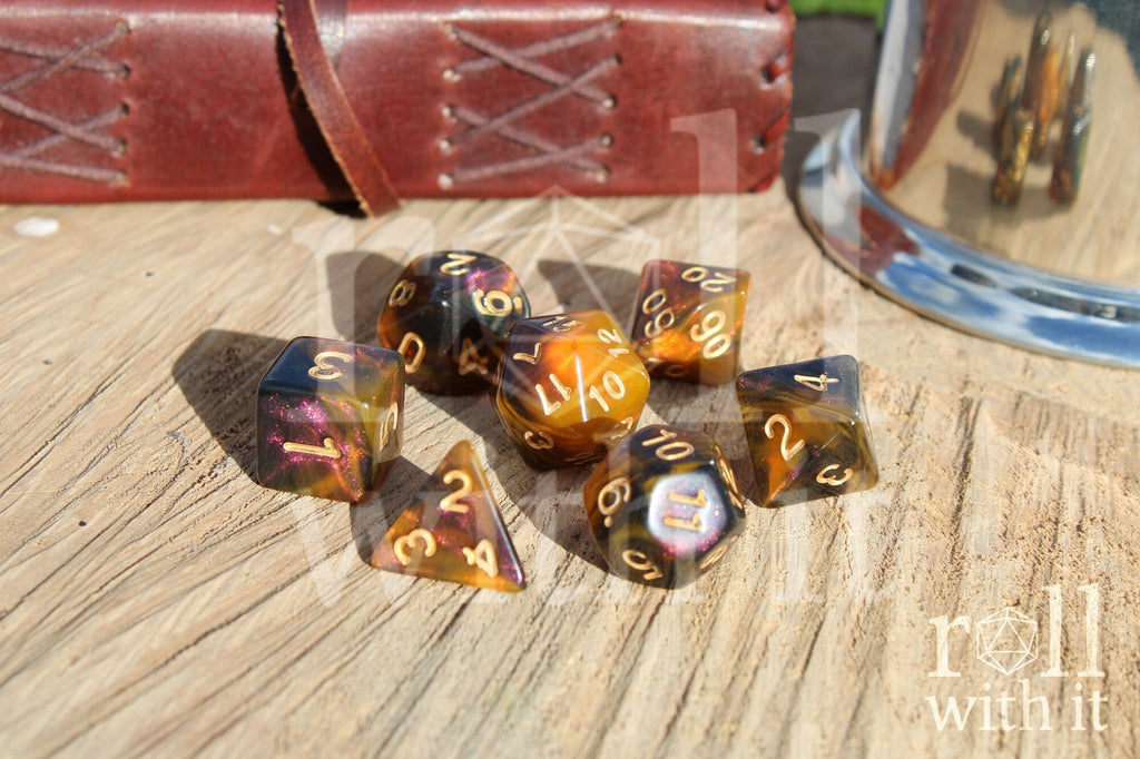 Set of 7 glittery pink, smoky amber and orange swirling polyhedral roleplaying dice on a wooden surface with a leather journal and shiny tankard in the background