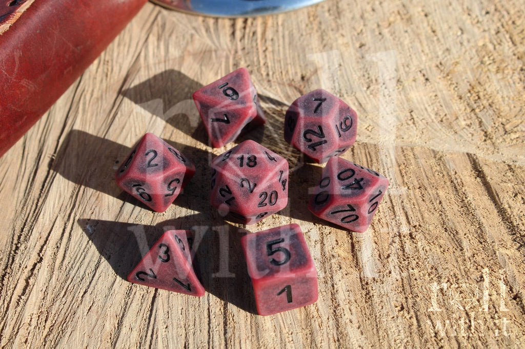 Matte red polyhedral roleplaying dice with a worn look with black lettering set on a wooden surface