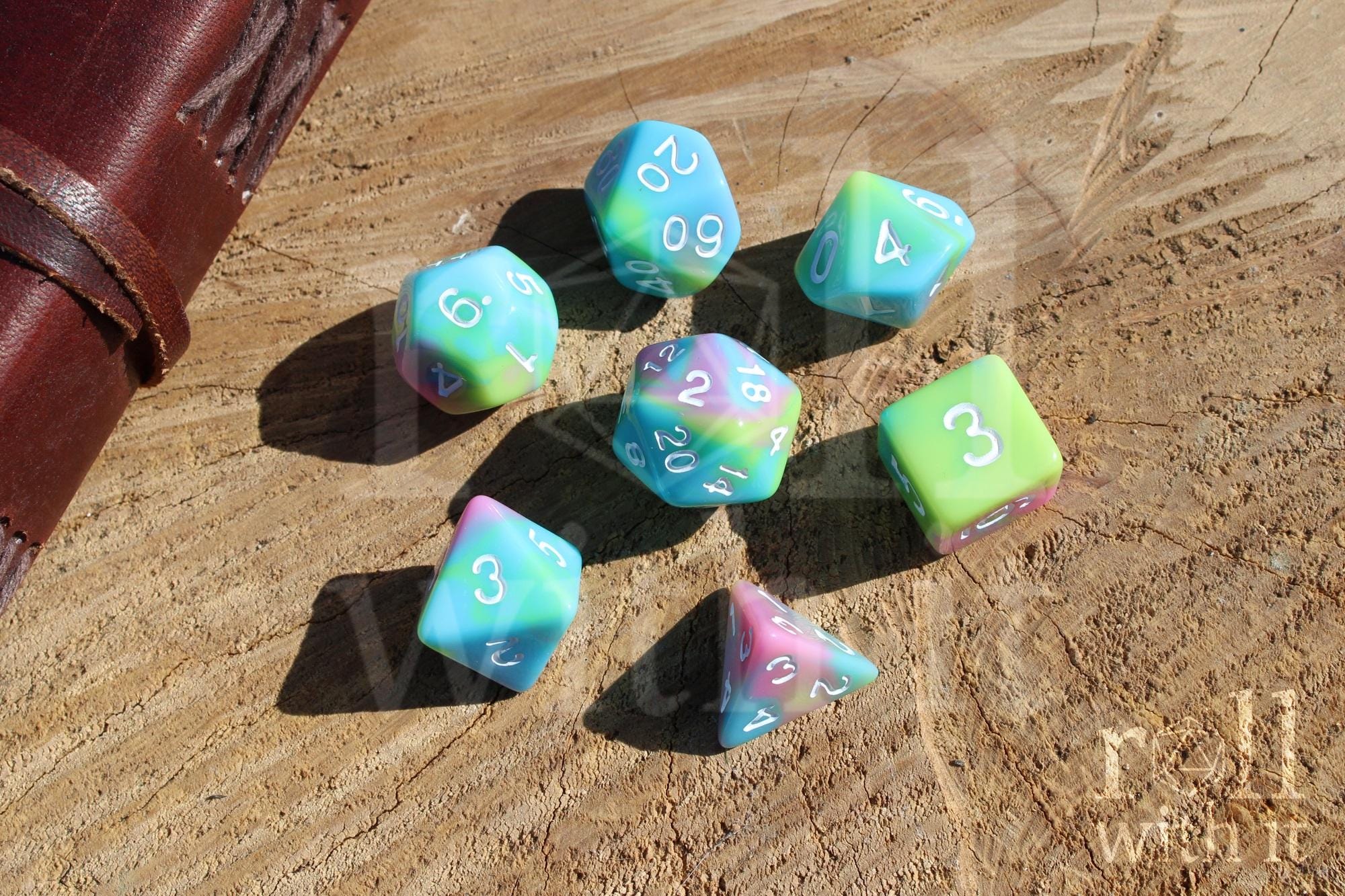 Set of colourful pastel rainbow polyhedral DND dice with white numbering on a wooden surface with a leather-bound  notebook in the background 