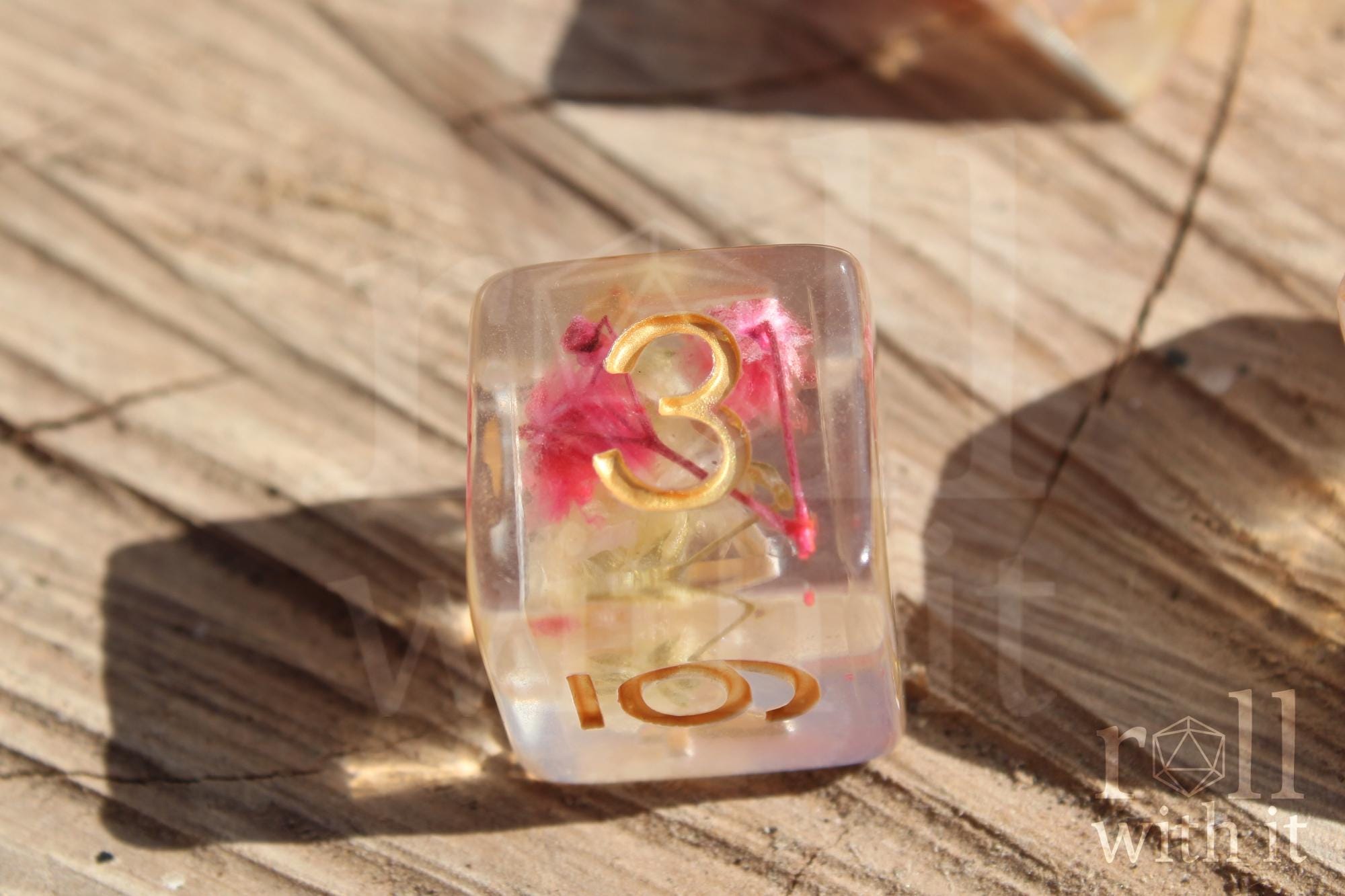 A close up of a transparent 6 sided dice with pink  yellow flowers inside with gold numbers on a wooden surface