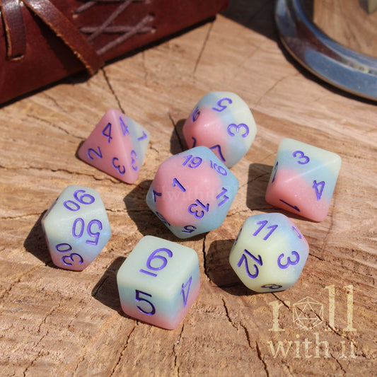 Set of pastel-colored pink blue yellow layered matte dice with purple numbers on a wooden surface, featuring 'roll with it' branding.