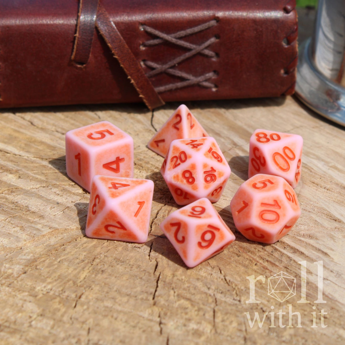 Set of pink matte worn polyhedral dice on a wooden surface with 'roll with it' branding.