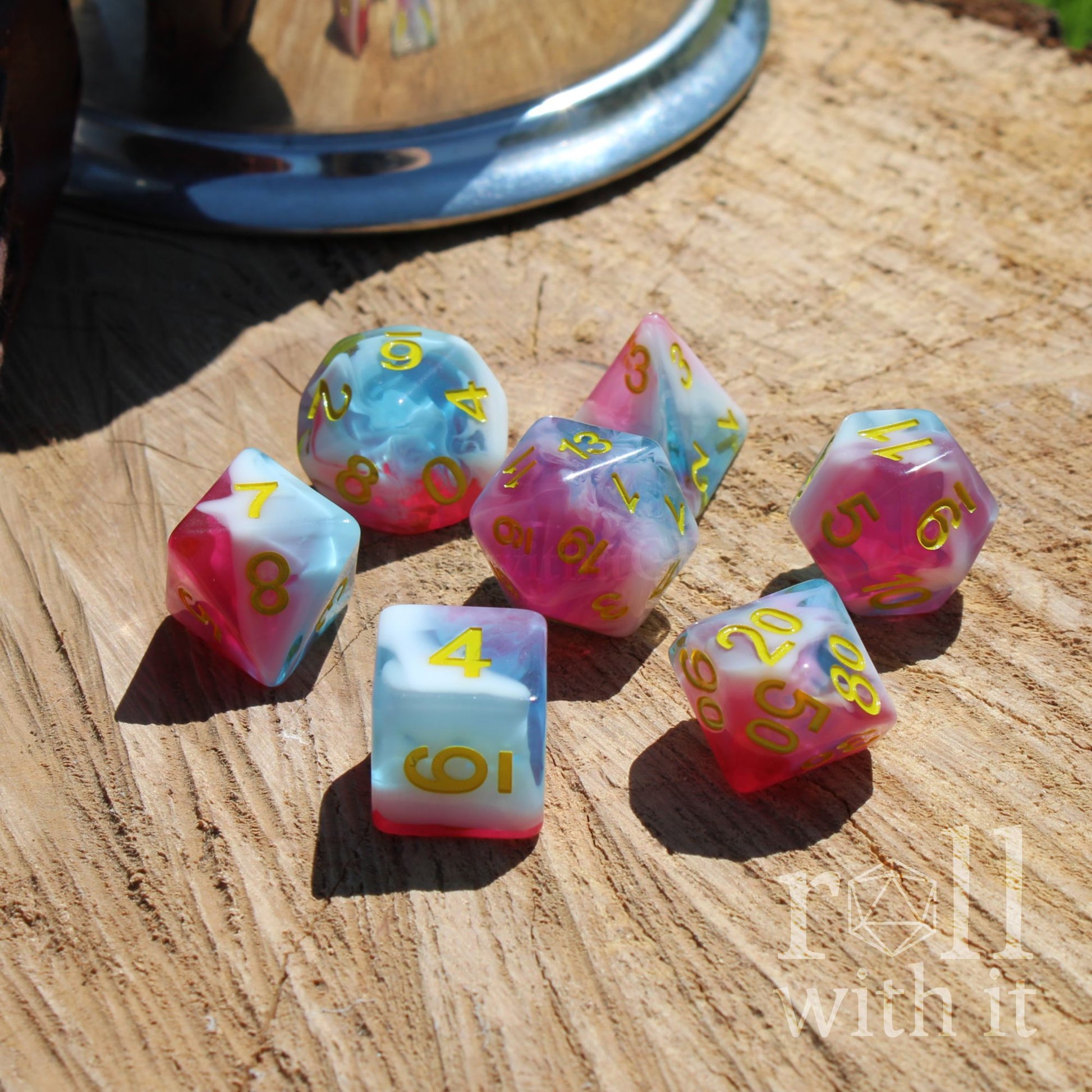 Set of colorful mixed swirling pink white blue polyhedral dice on a wooden surface with 'roll with it' text.