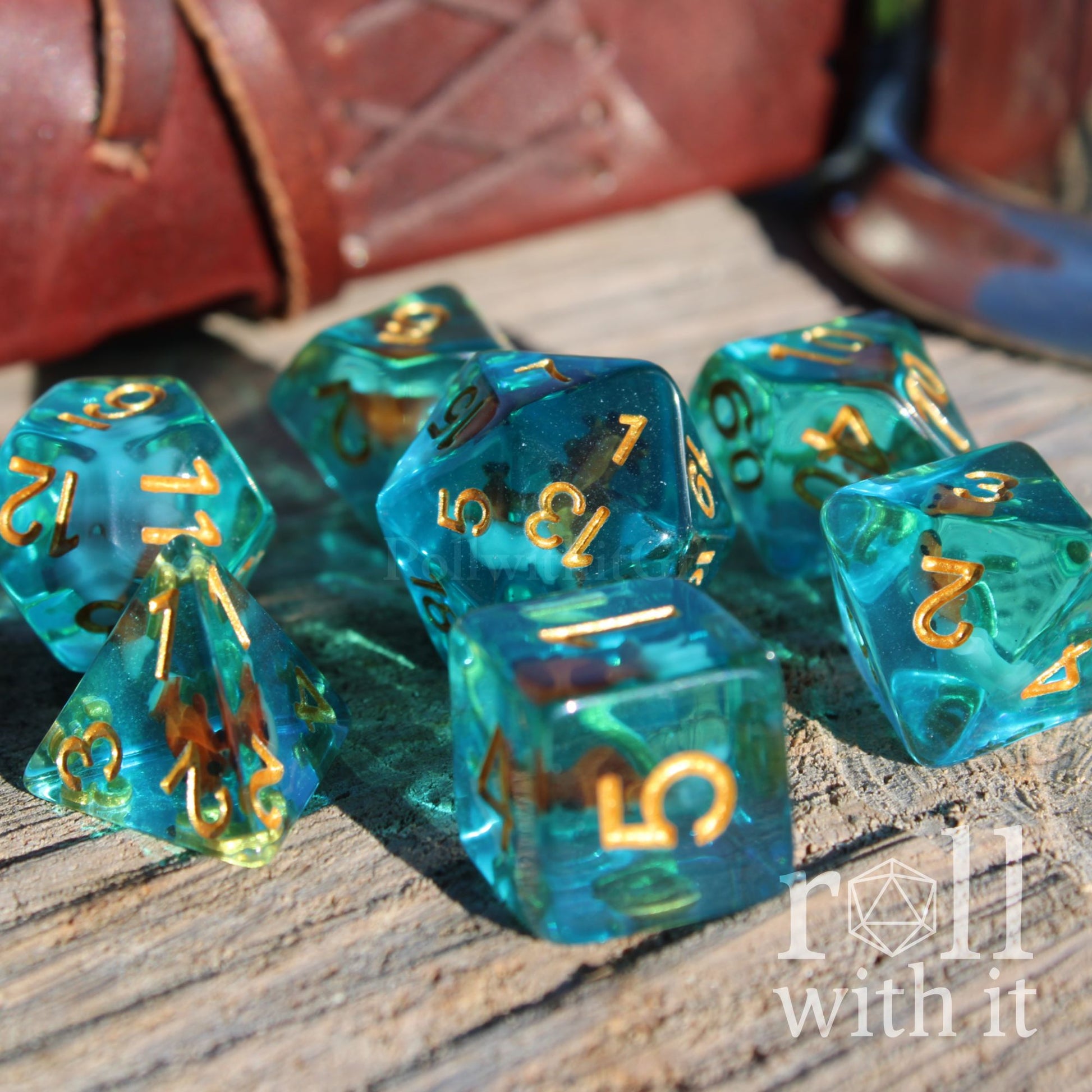 Set of teal and gold polyhedral dice with a miniature fish swimming inside on a wooden surface with 'roll with it' branding.